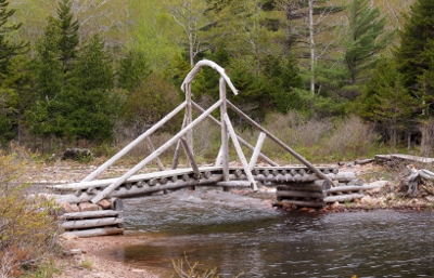 [Triangular trusses above a log-planked foot surface with an arched piece of wood across the walkway in the middle of the bridge.]